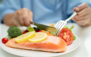 Person cutting a piece of cooked fish on a plate next to other foods rich in Omega 3’s.